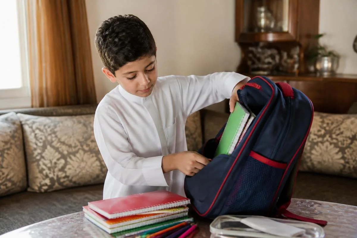 A young boy packing notebooks and pencils into his school backpack at home